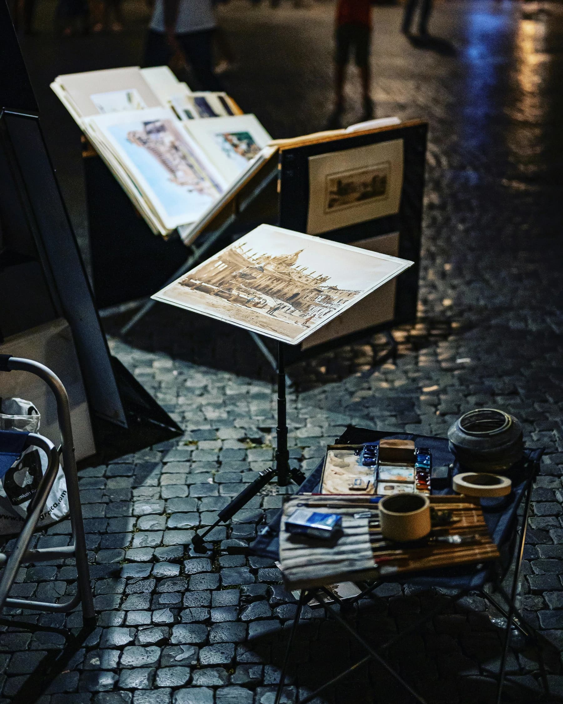 A street artist's watercolor setup on wet Roman cobblestones at night